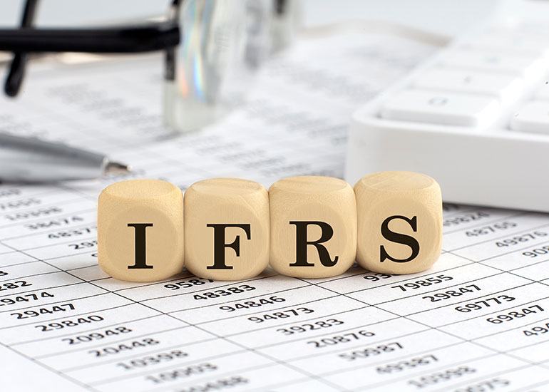 Auf den Würfeln steht „IFRS”. Jeder Buchstabe ist auf einem eigenen Würfel zu sehen. Unter den Würfeln liegt ein Zettel mit Zahlen. Im Hintergrund sind ein Kugelschreiber und eine Tastatur zu sehen.