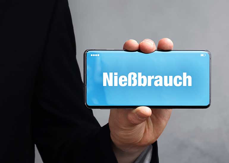 Eine Person hält ein Mobiltelefon in der Hand, auf dessen Display „Nießbrauch“ steht.