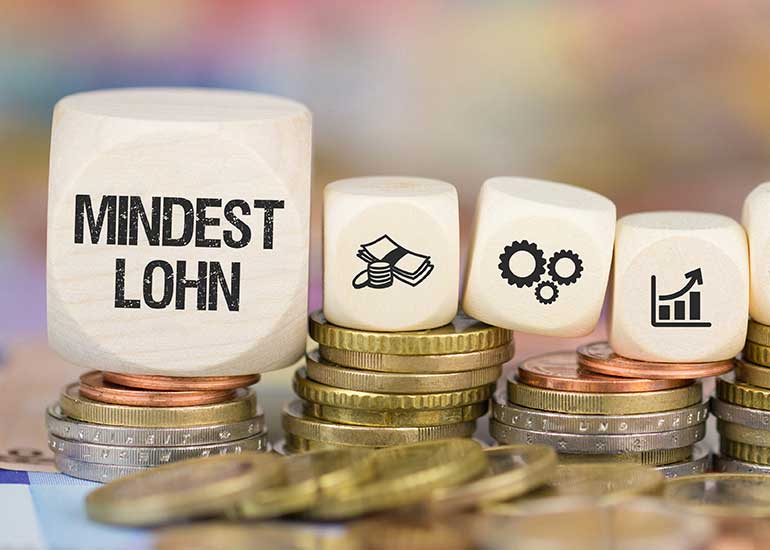 Auf einem Würfel steht „Mindestlohn“. Daneben liegen Würfel mit Steuersymbolen. Die Würfel liegen auf Münzstapeln.