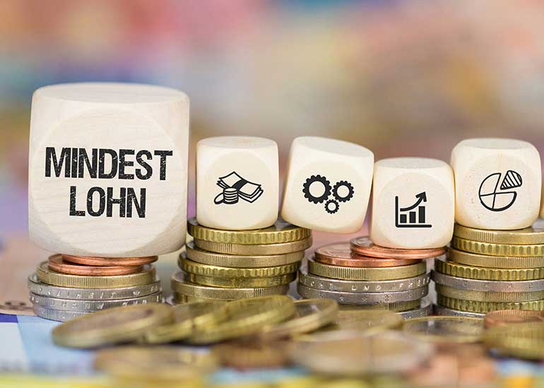 Auf einem Würfel steht „Mindestlohn“. Daneben liegen Würfel mit Steuersymbolen. Die Würfel liegen auf Münzstapeln.
