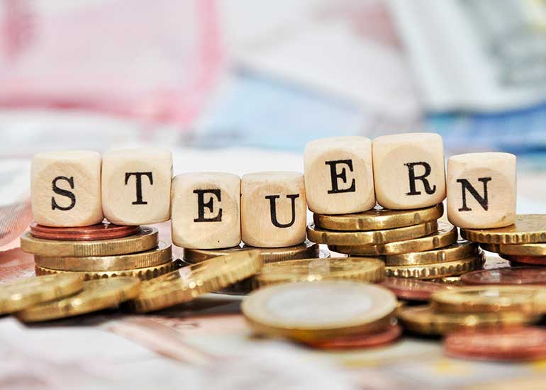 Der Begriff "Steuern" ist auf Würfeln geschrieben. Jeder Buchstabe ist auf einem einzelnen Würfel abgebildet. Alle Würfel stehen jeweils auf einem Turm aus Münzen.