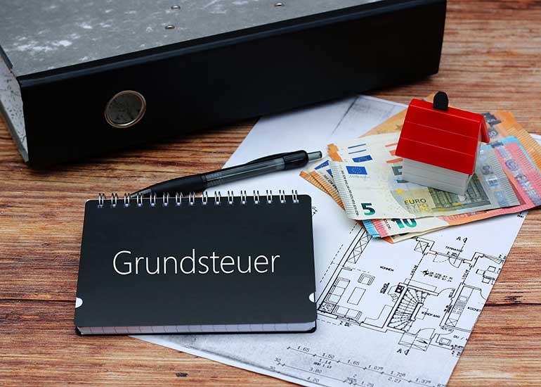 Auf dem Tisch liegt ein Bauplan. Darauf liegen Euro-Banknoten, ein Kugelschreiber, ein Aktenordner sowie ein Modell eines Hauses. Ebenso ein Notizbuch mit der Beschriftung „Grundsteuer”.