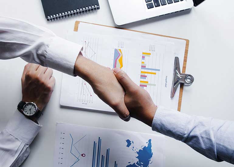 Über einem Schreibtisch, auf dem Blätter mit Grafiken liegen, schütteln sich zwei Personen die Hände.