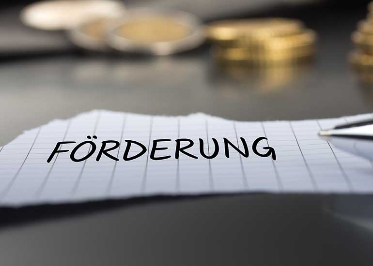 Auf einem Tisch liegt ein Zettel mit der Aufschrift „Förderung“. Im Hintergrund liegen Euro-Münzen.