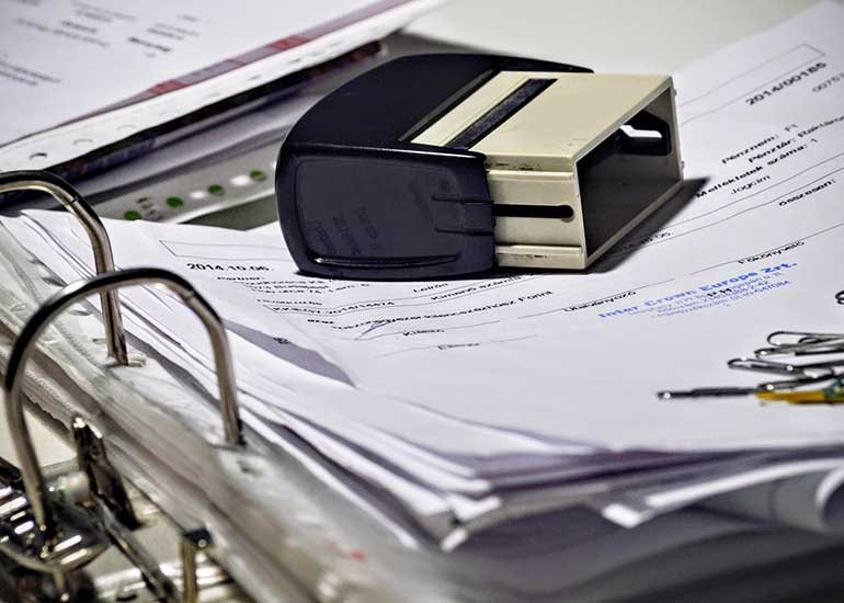 Auf einem Aktenordner, in dem viele Papiere abgeheftet sind, liegen ein Stempel und einige Büroklammern.