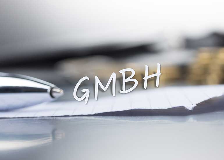 Die Bildcollage zeigt einen Tisch, auf dem ein Blatt Papier, ein Kugelschreiber und im Hintergrund Euro-Münzen liegen. Über dem Papier schwebt der Schriftzug „GmbH”.