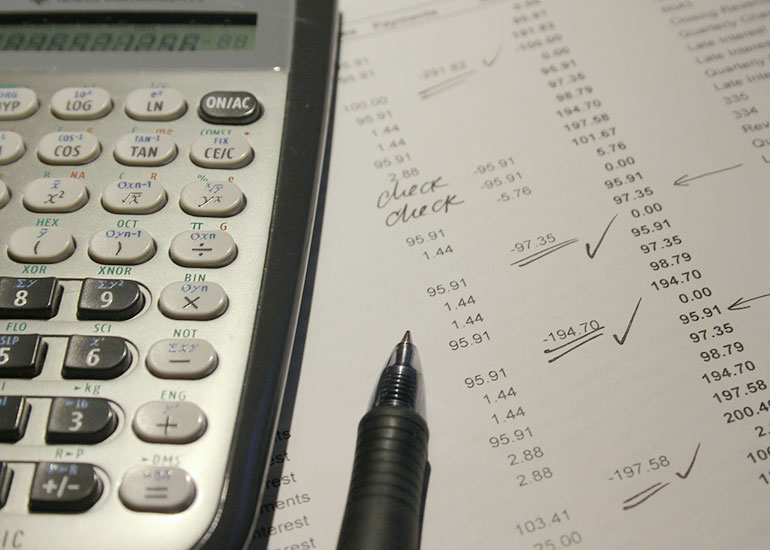 Neben einem Taschenrechner liegt ein Blatt Papier mit Zahlenspalten. Einzelne Zahlen wurden mit einem Stift als geprüft markiert. Der Stift liegt auf dem Papier. 
