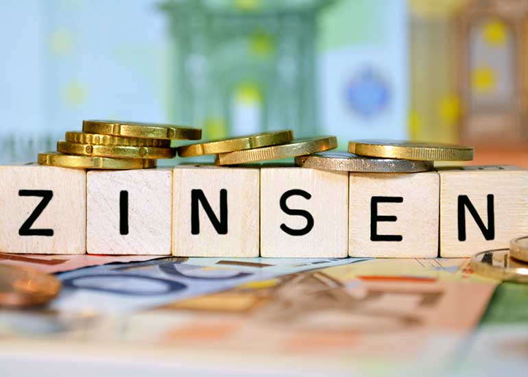 Der Begriff „Zinsen” ist aus Holzklötzen geschrieben. Jeder Buchstabe steht auf einem eigenen Klotz. Darauf liegen Münzen und darunter sind Euro-Banknoten zu erkennen.