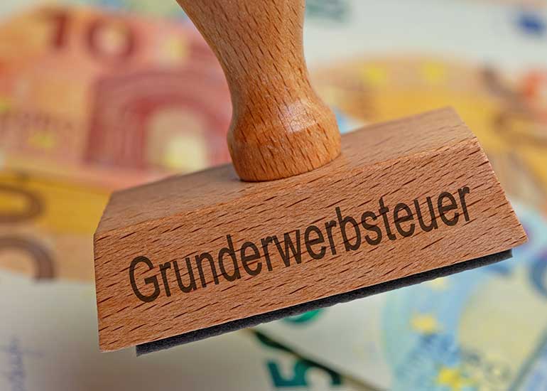Auf dem Stempel steht der Begriff „Grunderwerbsteuer”. Im Hintergrund sind Euro-Banknoten zu erkennen.