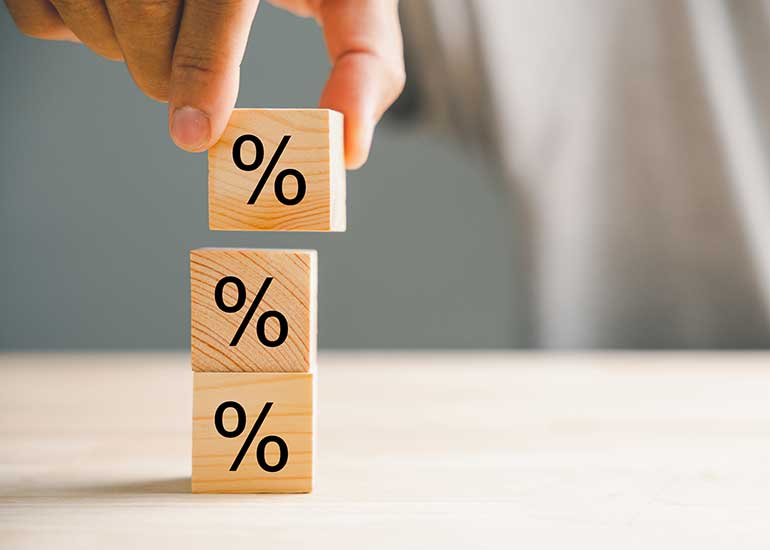 Eine Person stellt drei Holzklötzchen auf einen Tisch. Auf allen drei Klötzchen ist ein Prozentzeichen abgebildet.