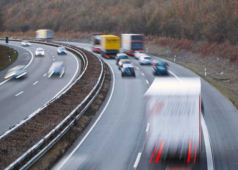 Mehrspurige Autobahn in einer Kurve mit dichtem Verkehr; mehrere Autos und Lastwagen fahren in beide Richtungen, einige davon verwischt durch Bewegung. Im Hintergrund stehen herbstliche Bäume am Hang.
