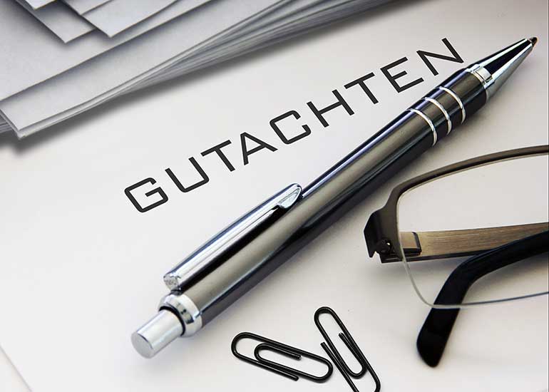 Auf dem Tisch liegen Papiere. Auf einem davon steht der Text „Gutachten”. Daneben sind ein Kugelschreiber, Büroklammern und eine Lesebrille zu sehen.