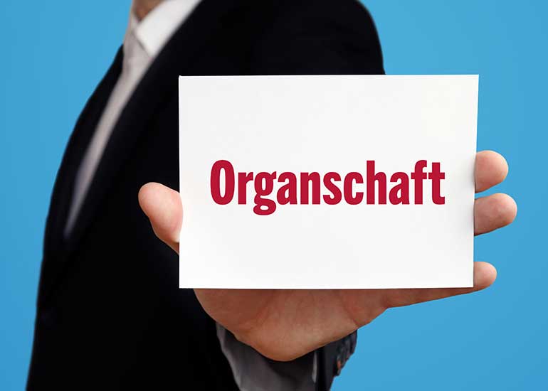 Eine Person mit Anzug hält einen Zettel mit der Aufschrift „Organschaft” in die Höhe.