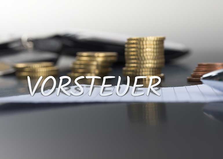 In einer Bildcollage ist der Schriftzug „Vorsteuer” vor einem Tisch platziert, auf dem ein Blatt Papier und ein Stapel Euromünzen liegen.
