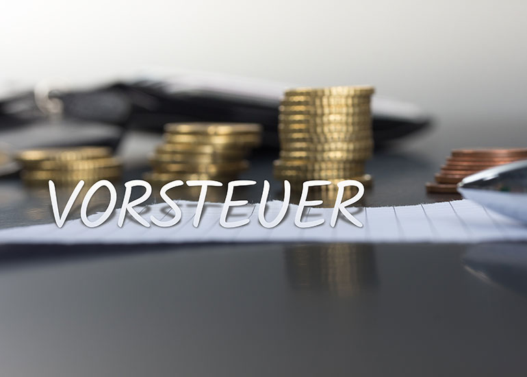 In einer Bildcollage ist der Schriftzug „Vorsteuer” vor einem Tisch platziert, auf dem ein Blatt Papier und ein Stapel Euromünzen liegen.