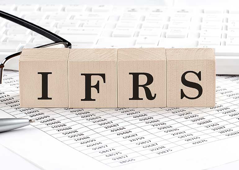 Auf einem Blatt mit Zahlentabellen steht eine Tastatur, eine Brille und ein Kugelschreiber. Zudem steht der Begriff „IFRS” auf Holzklötzchen. Jeder Buchstabe steht auf einem Klotz.