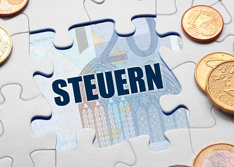 Aus einem Puzzle fehlt ein Teil. Darunter ist der Begriff „Steuern” zu lesen und eine Euro-Banknote zu erkennen. Daneben liegen Euro-Münzen.