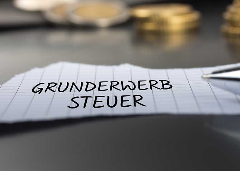 Auf einem Tisch liegt ein Zettel, auf dem „Grunderwerbsteuer” steht. Im Hintergrund sind Stapel aus Münzen und daneben ein Kugelschreiber zu sehen.