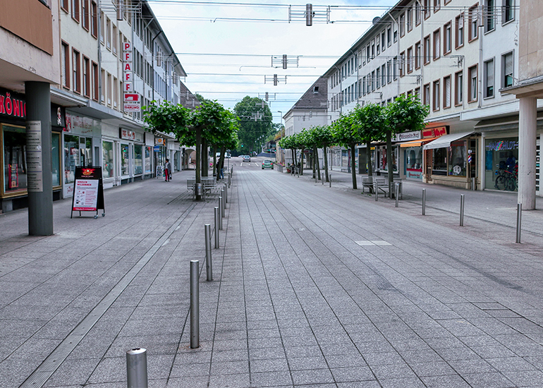 Breite, nahezu menschenleere Fußgängerzone in einer Stadt. Moderne Geschäftsgebäude und Schaufenster säumen beide Seiten der gepflasterten Straße. Gleichmäßig angeordnete Bäume, Sitzgelegenheiten und Poller strukturieren den öffentlichen Raum. Die Perspektive führt entlang der Einkaufsstraße in die Ferne unter einem leicht bewölkten Himmel.