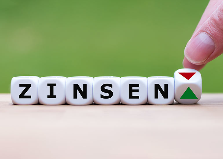 Das Wort „Zinsen” ist aus Würfeln geschrieben. Jeder Buchstabe steht auf einem eigenen Würfel. Eine Hand dreht einen weiteren Würfel um, sodass statt eines Pfeils nach oben ein Pfeil nach unten erscheint.