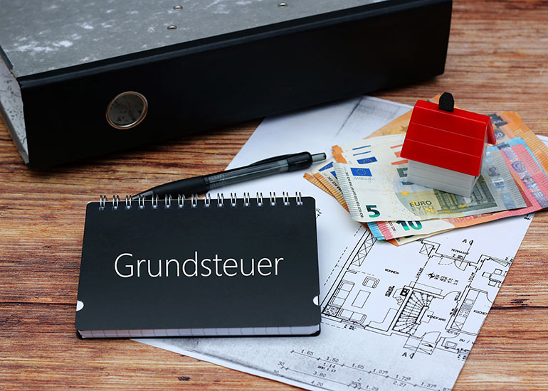 Auf dem Tisch liegt ein Bauplan. Darauf liegen Euro-Banknoten, ein Kugelschreiber, ein Aktenordner sowie ein Modell eines Hauses. Ebenso ein Notizbuch mit der Beschriftung „Grundsteuer”.