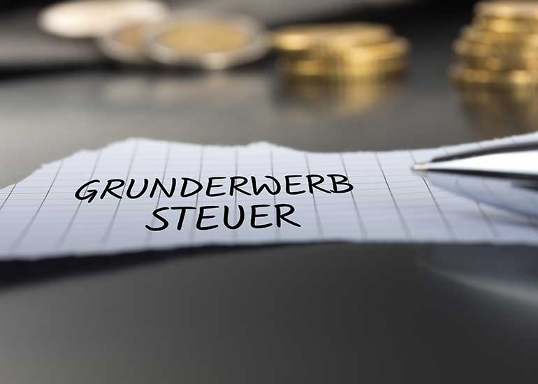 Auf einem Tisch liegt ein Zettel, auf dem „Grunderwerbsteuer” steht. Im Hintergrund sind Stapel aus Münzen und daneben ein Kugelschreiber zu sehen.