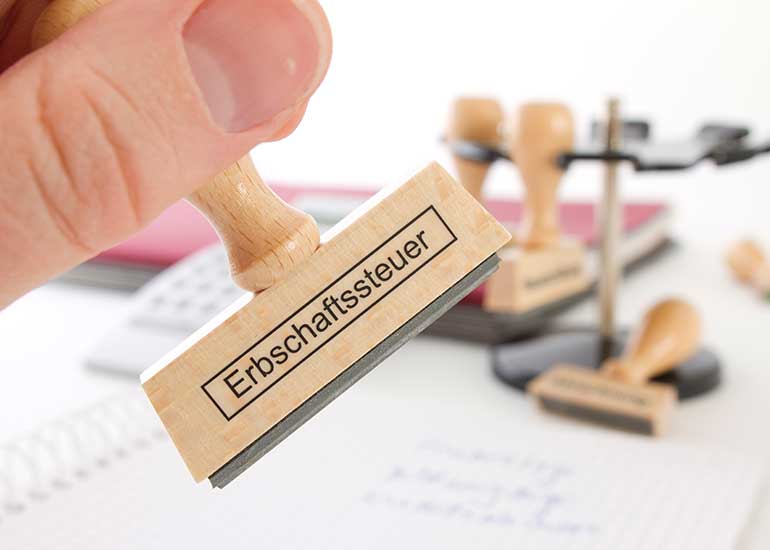 Eine Hand hält einen Stempel mit der Aufschrift "Erbschaftssteuer". Im Hintergrund liegen weitere Stempel auf einem Schreibtisch.