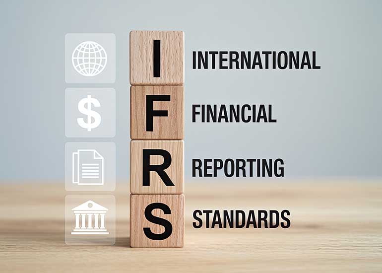 Die Bildcollage zeigt vier übereinandergestapelte Holzbausteine, die das Akronym „IFRS“ bilden. Daneben steht die ausgeschriebene Bezeichnung „International Financial Reporting Standards“. Ergänzende Symbole für globale Finanzberichterstattung, Dokumentation und Regulierung verdeutlichen den Bezug zu internationalen Rechnungslegungsstandards. Das Bild wurde mithilfe von KI erstellt.