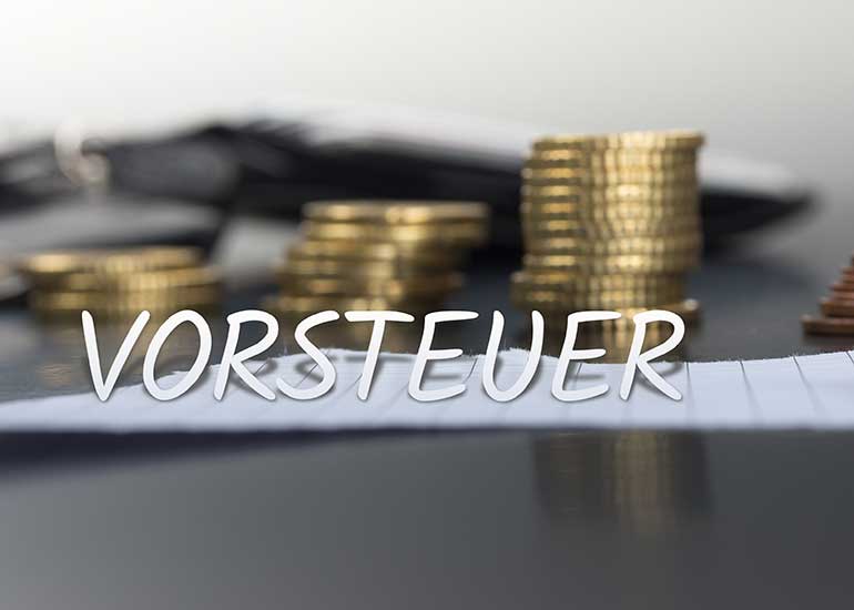 In einer Bildcollage ist der Schriftzug „Vorsteuer” vor einem Tisch platziert, auf dem ein Blatt Papier und ein Stapel Euromünzen liegen.