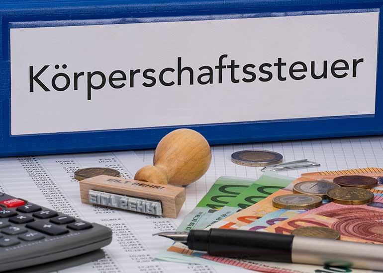 Aktenordner mit der Aufschrift „Körperschaftssteuer“ auf einem Schreibtisch mit Euro-Banknoten, Münzen, Taschenrechner, Stempel und Stift.