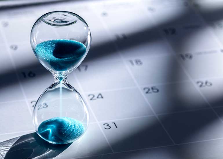 Sanduhr mit blauem Sand vor einem unscharfen Kalenderblatt. Das Bild symbolisiert Fristen, Termine oder Zeitabläufe.