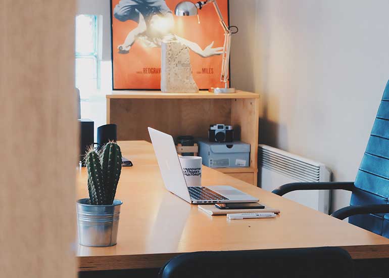 Arbeitsplatz in einem Büro mit Schreibtisch, geöffnetem Laptop, Bürostuhl und Dekoration. Im Hintergrund befinden sich Regale, eine Schreibtischlampe und ein Wandbild.