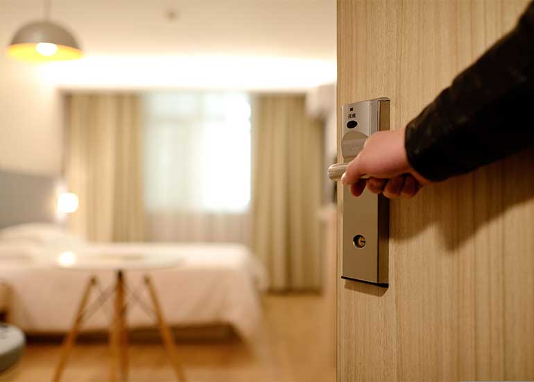 Hand öffnet die Tür zu einem Hotelzimmer mit Bett und Sitzgelegenheit.