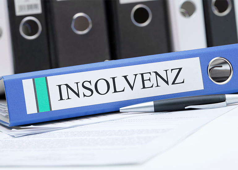 Blauer Aktenordner mit der Aufschrift ‚Insolvenz‘ auf einem Schreibtisch. Im Hintergrund sind weitere Ordner und Dokumente zu sehen.