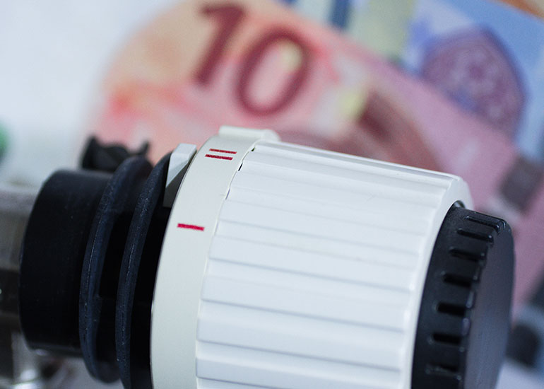 Nahaufnahme eines Heizkörperthermostats. Im Hintergrund sind unscharf Euro-Scheine zu sehen, die als Symbol für Energiekosten und Heizkosten stehen.